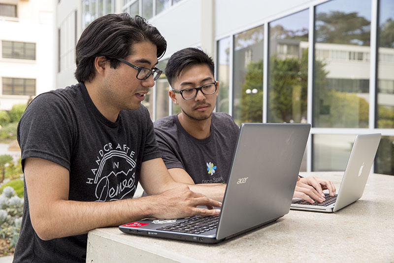 A male student with glasses sits outside the library. His laptop is open in front of him, and another male student with glasses on is sitting next to him. They are looking at the laptop screen.