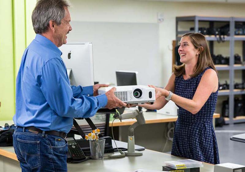 A smiling woman in a blue dress, standing behind a service counter, hands a white projector unit to a man in a blue shirt.