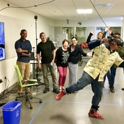 a crowd gathers around a person using a virtual reality headset with their limbs splayed out in an active motion